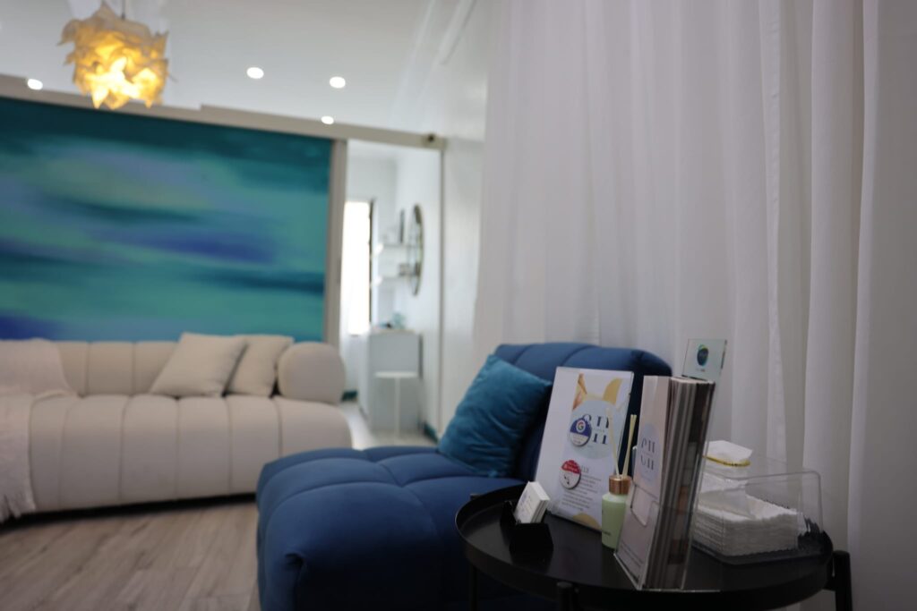 Luxurious clinic waiting room interior at The Contour Hub showing a plush deep blue daybed lounge with branded clinical brochures and Google review stand on a side table next to sheer white privacy curtains.